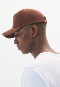 Brown baseball cap with a curved brim and stitched eyelets, featuring a logo on the front panel. Worn at an angle, showing a textured surface.