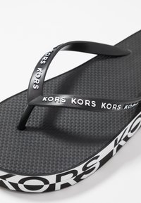 Musta sandaali, jossa on teksturoitu pohja, tyylikkäät mustat hihnat, joissa on toistuva valkoinen "KORS"-logo, sekä kontrastina valkoinen raita pohjassa.