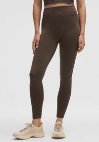 Leggings bruns avec une taille haute, dotés de coutures sculptées et d'une texture lisse et extensible. Associés à des chaussures de sport de couleur claire.