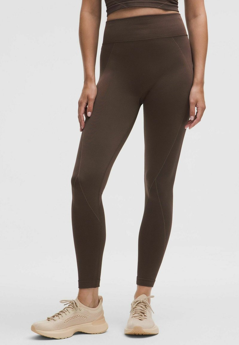 Leggings bruns avec une taille haute, dotés de coutures sculptées et d'une texture lisse et extensible. Associés à des chaussures de sport de couleur claire.