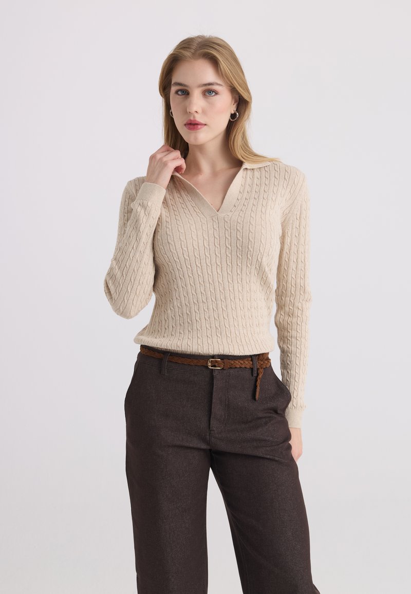 Femme blonde portant un pull beige en maille torsadée avec col en V et un pantalon marron foncé avec ceinture tressée marron, debout devant un fond uni.