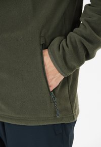 Chaqueta de forro polar verde oliva con un bolsillo con cremallera, tela texturizada y puños ajustados. Tirador de cremallera visible y ribete en contraste.