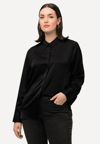 Camicia nera di satin a maniche lunghe con colletto classico e chiusura con bottoni. Indossata con jeans scuri, che mettono in risalto una texture morbida e una vestibilità rilassata.