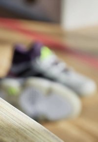 Grå sportssko med lilla accenter og neongrønne detaljer. Teksturerede såler, snørebånd synlige, let sløret baggrund af trægulv.