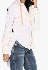 Veste zippée légère et irisée avec des cordons orange, des manches longues, des fentes latérales et un design épuré. Associée à un denim déchiré.