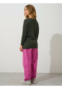 Dunkelgrüner Langarmpullover mit lässigem Schnitt, kombiniert mit lockeren rosafarbenen Hosen und grauen Sneakers, vor einem schlichten Hintergrund.