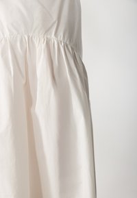 Robe beige clair en tissu doux, présentant une taille froncée et une jupe fluide, avec une texture lisse et un éclat subtil.
