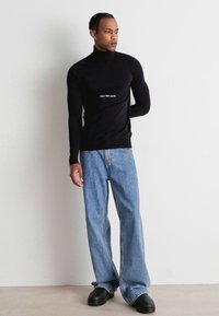 Maglietta nera a maniche lunghe con collo alto e logo bianco, abbinata a jeans a gamba larga azzurri e scarpe nere con suola chunky.