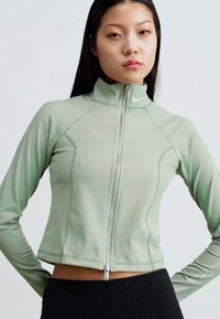 Pepperminta zaļa, pieguļošs, ar rāvējslēdzēju aiztaisāms žakets ar augstu apkakli un kontrastējošiem šuvēm. Tajā ir Nike logo pie kakla un ribota melna auduma apakšdaļa.