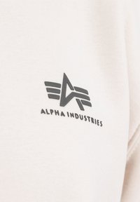 Witte sweater met een zwart logo. Het logo bevat gestileerde vleugels en de tekst "ALPHA INDUSTRIES" eronder. Gladde stoftextuur.