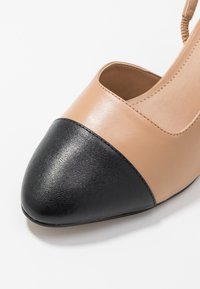 Beige leather shoe with a black textured toe cap. Features an open side design and smooth finish, showcasing contrasting color elements.