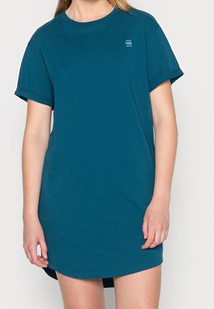 Personne portant une robe t-shirt manches courtes bleu sarcelle avec un petit logo sur la poitrine gauche, debout devant un fond uni.