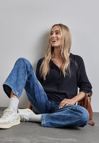Maglioncino blu con bottoni, jeans blu larghi, sneaker bianche, calzini grigi, borsa marrone, seduto su un pavimento grigio contro un muro chiaro.
