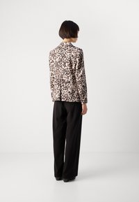 Persoon met kort haar, gekleed in een beige jassen met luipaardprint en zwarte wijde broek, staande met de rug naar ons toe tegen een plain witte achtergrond.