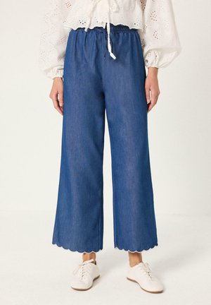 Mujer que lleva pantalones vaqueros azules de tiro alto y pierna ancha con bajos festoneados, combinados con zapatillas blancas y una blusa blanca con ojales.