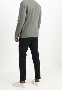 Mężczyzna w jasnoszarym blezerze, czarnych zwężających się spodniach i białych trampkach, stoi plecami do obiektywu na jednolitym białym tle.