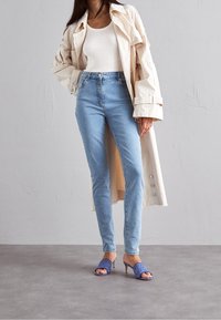 Cappotto trench beige sopra una canotta bianca a costine, jeans azzurri a vita alta e sandali con tacco a fantasia blu. Texture morbide in evidenza.