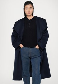Manteau en mélange de laine bleu marine avec grands revers, superposé à un pull en maille noir et un jean en denim bleu, mettant en avant une coupe décontractée et des lignes épurées.