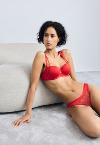 Set de lenjerie din dantelă roșie, care include un sutien cu cupe modelate și bretele decorative, completat cu chiloți din dantelă transparentă asortați. Texturi de fundal moi.