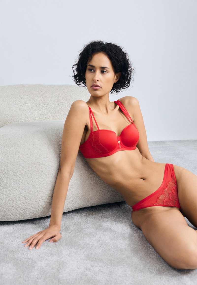 Set de lenjerie din dantelă roșie, care include un sutien cu cupe modelate și bretele decorative, completat cu chiloți din dantelă transparentă asortați. Texturi de fundal moi.