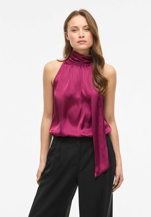 Femme portant un haut sans manches en satin magenta avec un col montant noué, associé à un pantalon noir sur mesure, sur fond blanc.