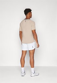 Beige Sporthemd mit kurzen Ärmeln, Rundhalsausschnitt und einem dezenten Muster auf dem Rücken, kombiniert mit weißen Shorts mit kleinen navyfarbenen Akzenten.