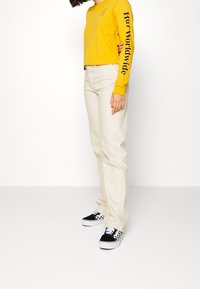 Gul långärmad skjorta med svart text på ärmen, kombinerad med krämfärgade byxor och svart-vit rutiga sneakers.