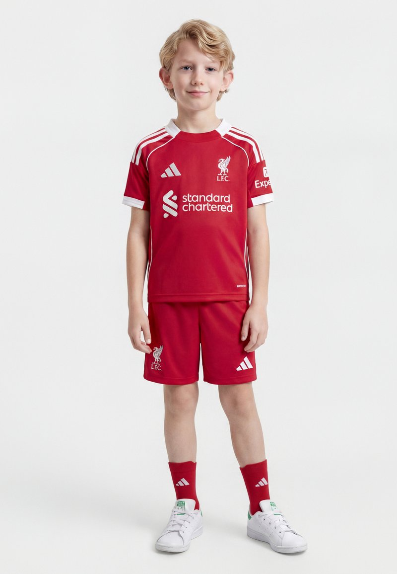 Blond jongetje staand, gekleed in het rode Liverpool FC voetbaltenue met witte Adidas-logo's, rode sokken en witte sneakers, tegen een witte achtergrond.