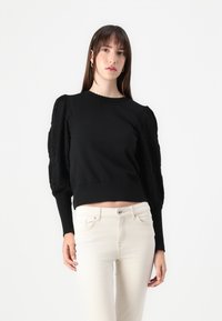 Pull nero con maniche puff e texture. Presenta un collo scallop e un orlo aderente. Abbinato a jeans chiari.