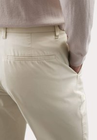 Beige Hosen aus glattem Stoff mit einer Gesäßtasche und einer schlanken Passform. Der Bund verfügt über Gürtelschlaufen und ist mit einem lockeren oberen Teil gestaltet.