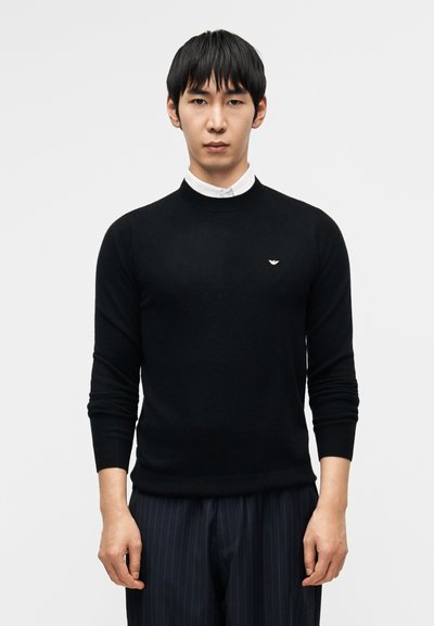 Pull noir en tricot avec un col rond, une coupe slim et des poignets côtelés, doté d'un petit logo sur la poitrine. Associé à un pantalon à fines rayures.