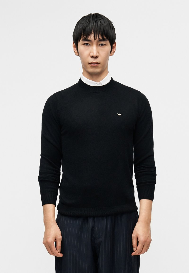 Μαύρο πλεκτό πουλόβερ με στρογγυλή λαιμόκοψη, εφ αρμοστό και με ριμπ μανσέτες, με ένα μικρό λογότυπο στο στήθος. Συνδυάζεται με παντελόνια με λεπτές γραμμές.