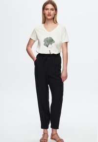 Hvid V-hals t-shirt med grøn bladmønster, parret med sorte tapered bukser med elastisk talje og manchetter, båret med brune sandaler.