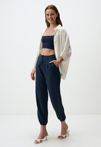 Weißes übergroßes Hemd mit aufgerollten Ärmeln über einem dunkelblauen Crop-Top. Marineblaue, plissierte Hosen in Knöchelhöhe und weiße Absatzsandalen mit offener Zehenpartie.