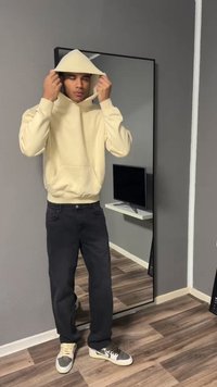 Jeune homme ajustant la capuche d'un sweat à capuche beige, debout devant un grand miroir dans une pièce minimaliste avec un sol en bois et une télévision murale.