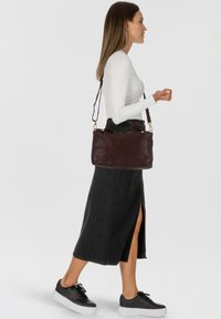 Bruine leren handtas met een ritsluiting, gouden hardware, dubbele handvatten en een afneembare schouderriem, gedragen door een persoon in een witte blouse en rok.