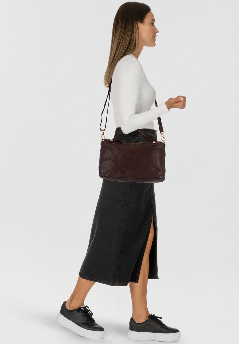 Bruine leren handtas met een ritsluiting, gouden hardware, dubbele handvatten en een afneembare schouderriem, gedragen door een persoon in een witte blouse en rok.