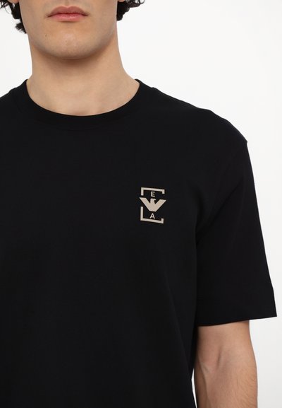 Juodas medvilnės marškinėlis su mažu smėlio spalvos logotipo dizainu kairiajame viršutiniame kampe, apvalia iškirpte ir trumpomis rankovėmis.