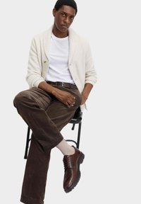 Eine cremefarbene Strickjacke über einem weißen T-Shirt, kombiniert mit braunen Cordhosen und braunen Lederschuhen, sitzt auf einem schwarzen Stuhl.