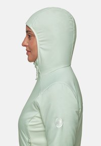 Femme en veste vert clair avec capuche ajustée zippée, profile lat�éral visible, et patch logo sur la manche supérieure sur fond uni.