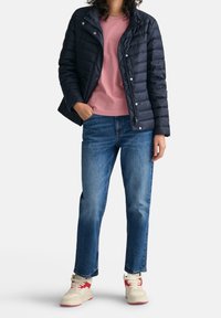 Person in einer marineblauen Steppjacke über einem rosa Hemd, blauen Jeans und weißen Sneakern mit roten Akzenten, steht mit einer Hand in der Tasche.