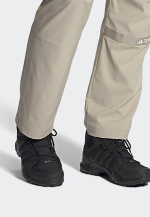 Czarne buty trekkingowe z teksturowanym materiałem i wieloma paskami, w połączeniu z jasnoszarymi spodniami z zapięciem na zatrzask u dołu.