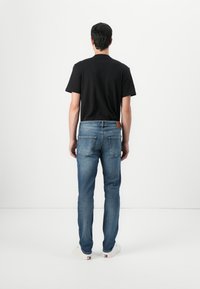 Black crew-neck t-shirt, blue denim jeans with a slim fit, visible back pockets, brown leather accent on waistband, white sneakers.
