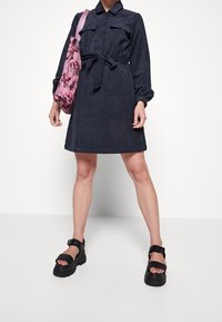 Abito camicia blu navy con cintura regolabile in vita e maniche lunghe, abbinato a una borsa floreale rosa e sandali neri con zeppa spessa. Texture liscia.