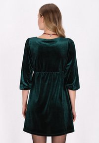 Rochie din catifea verde închis, cu maneci trei sferturi, talie strânsă și o textură netedă, având un decolteu rotund și o croială relaxată.
