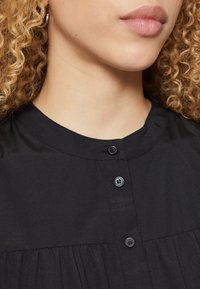 Black collared shirt with small buttons and pleated fabric details. Smooth texture and lightweight material, featuring a round neckline.