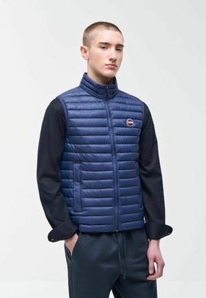 Mlad moški, ki nosi modro prešito brezrokavnik čez črno majico z dolgimi rokavi, roke ima v žepih temnih hlač, na enobarvnem ozadju.