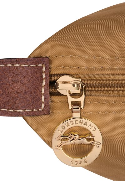 Brown leather zipper pull attached to a tan fabric bag, featuring a gold tag with a leaping horse logo and "LONGCHAMP 1948" inscription.