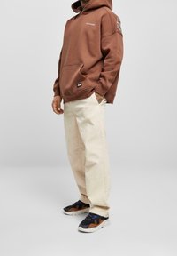 Sudadera marrón con capucha y un bolsillo delantero, combinada con pantalones de pierna ancha color beige claro y zapatillas negras con acentos en naranja y púrpura.