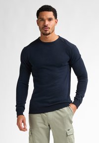 Mörkblå stickad långärmad tröja med rund halsringning, ribbade ärmslut och kant. Bärs med ljusa khaki cargo-byxor.